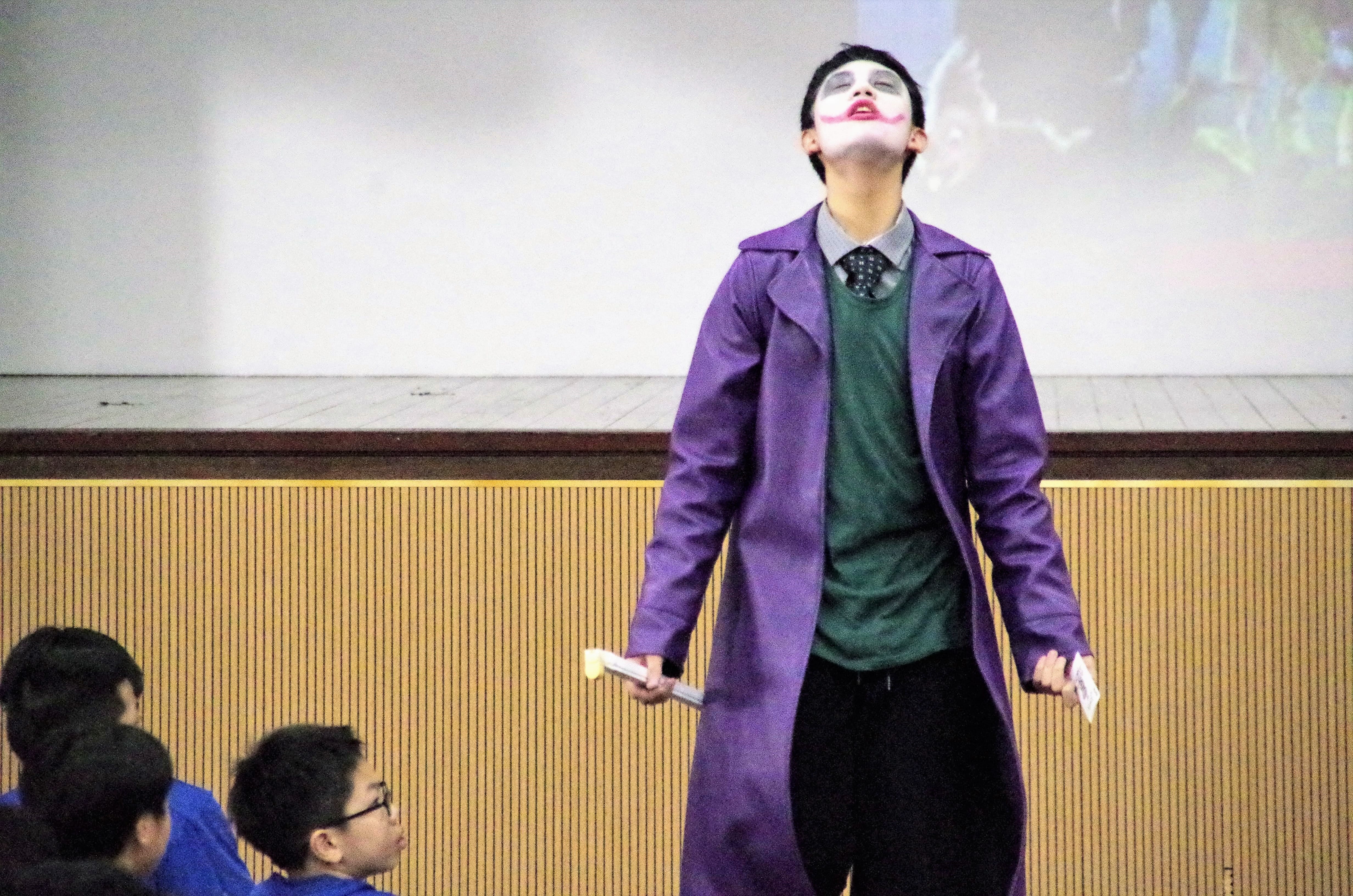 A student dressed as the Joker, in a purple coat and green shirt with white and red face paint, tilting his head back on stage.